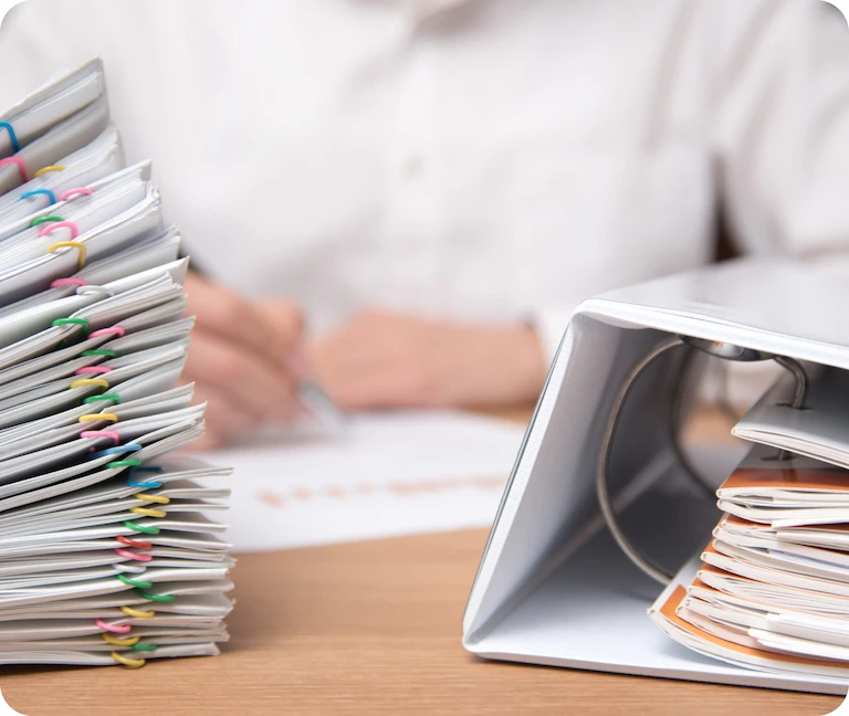 Stack of organized documents with colorful paper clips next to an open binder, with a person writing in the background