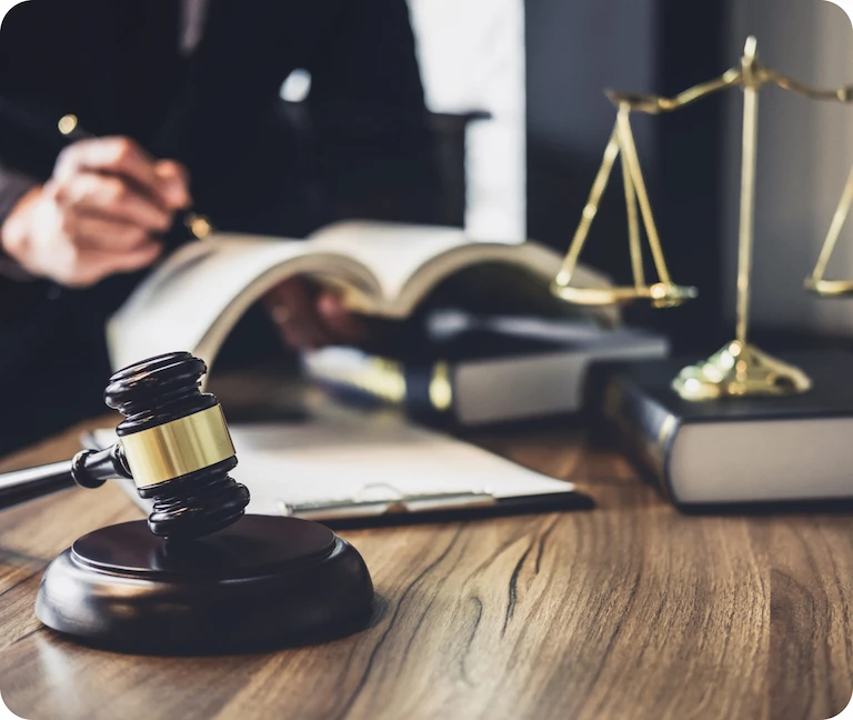 Wooden judge’s gavel on a desk with legal books, scales of justice, and a lawyer reviewing documents in the background